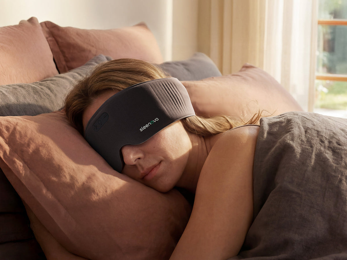 A woman sleeping soundly in bed with her eyes covered by a dark gray Eleveno sleep mask, surrounded by warm brown and gray pillows and a gray blanket.
